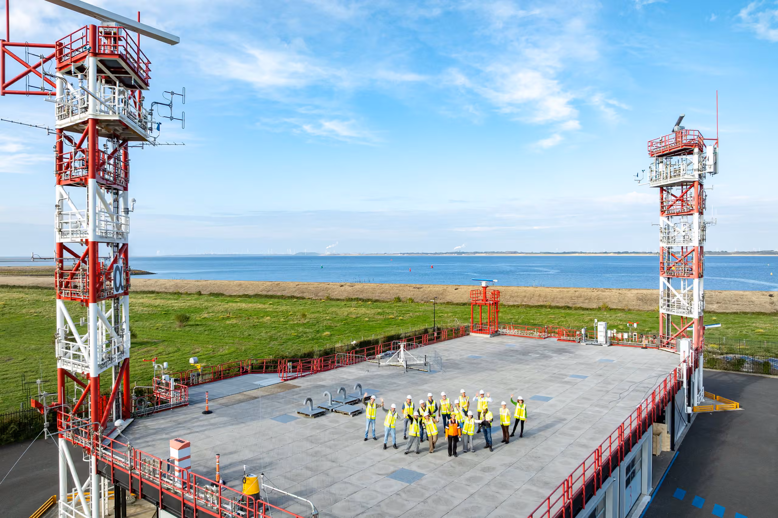 Een groep bezoekers met veiligheidsvesten en helmen poseert op de buitenopstelling van het Offshore Expertise Centrum, met een hoge rood-witte mast en het water op de achtergrond.