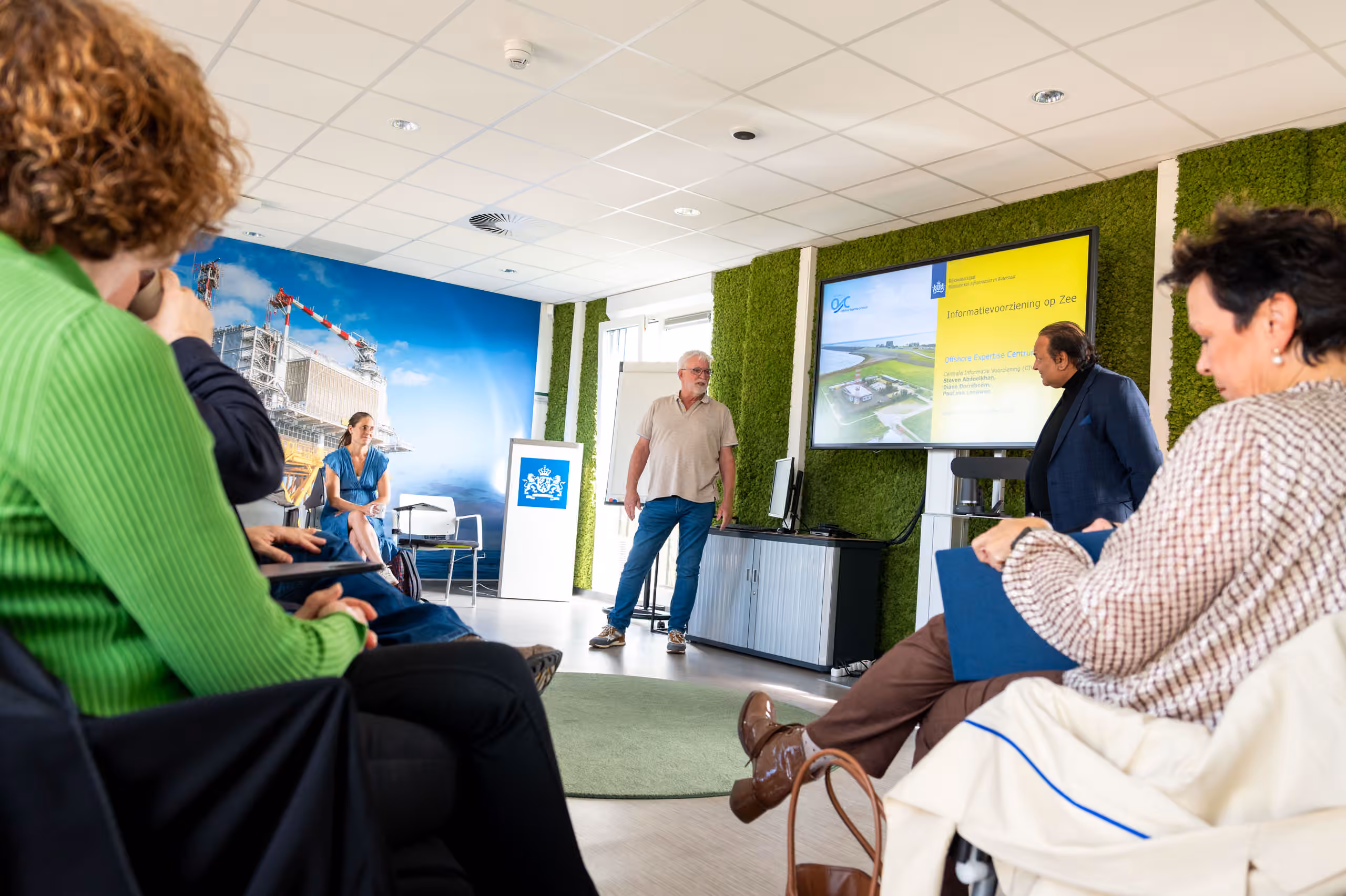 Bezoekers wonen binnen een presentatie bij over informatievoorziening op zee. Voorin staan twee sprekers bij een scherm met een presentatie over het Offshore Expertise Centrum.