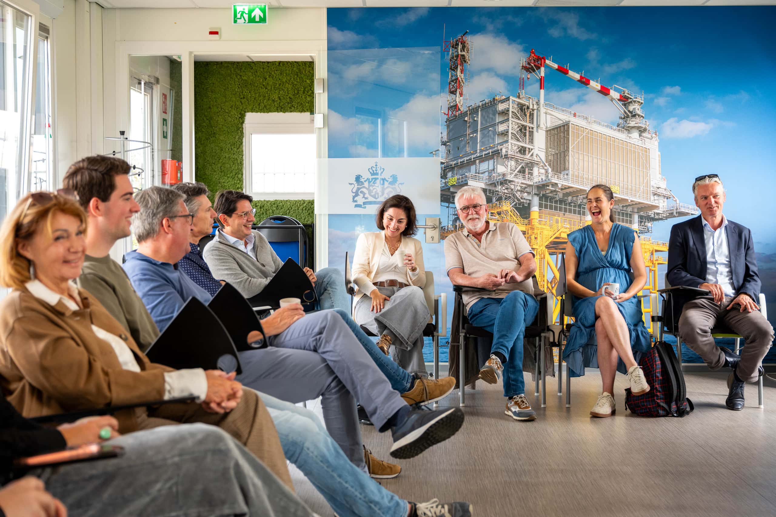 Een groep bezoekers zit in een kring tijdens een bijeenkomst in het Offshore Expertise Centrum. Op de achtergrond is een grote afbeelding van een offshoreplatform te zien.