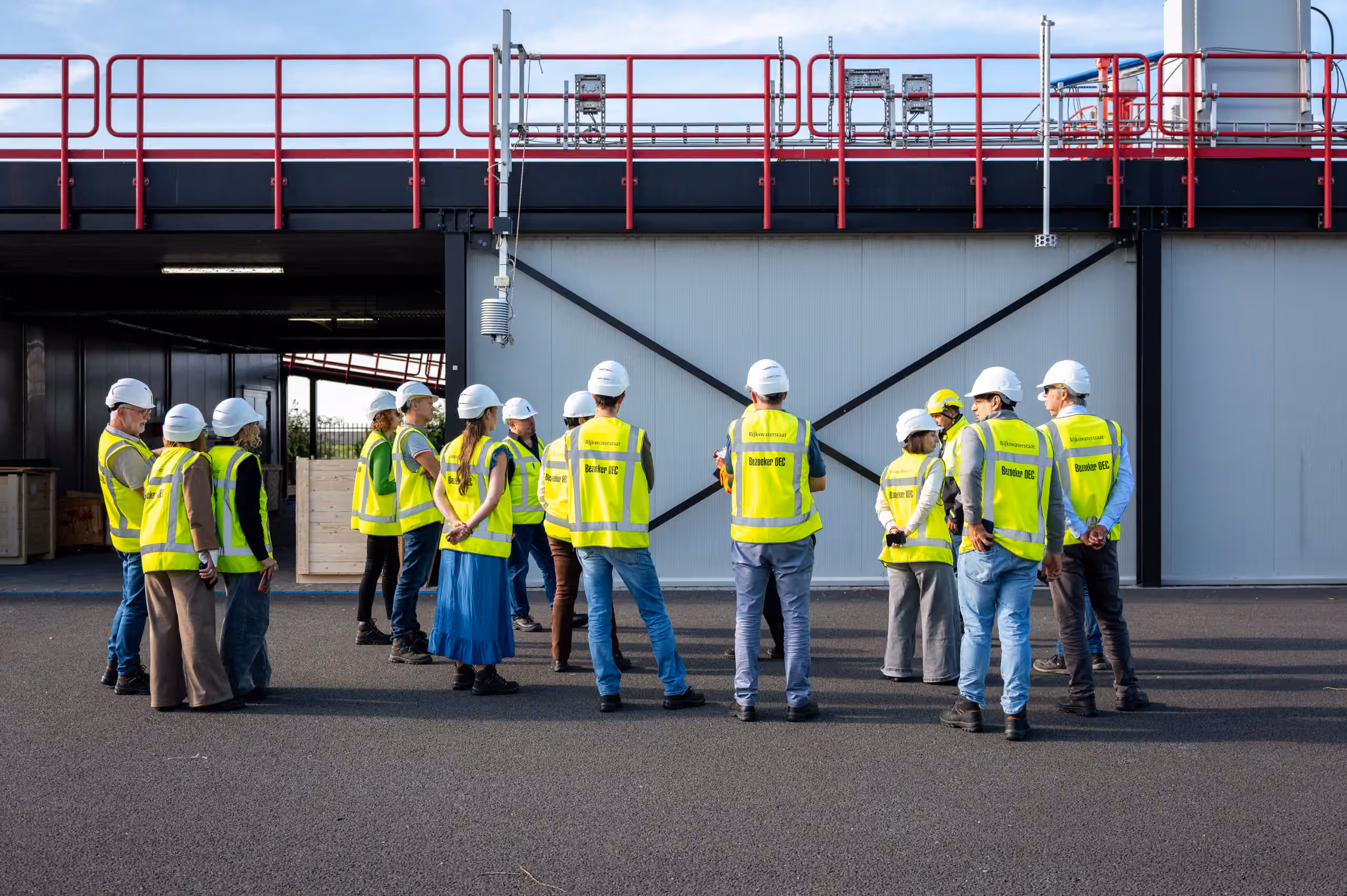 Een groep bezoekers met veiligheidsvesten en helmen staat bij de buitenopstelling van het Offshore Expertise Centrum, waar verschillende meetinstrumenten en sensoren zijn opgesteld.