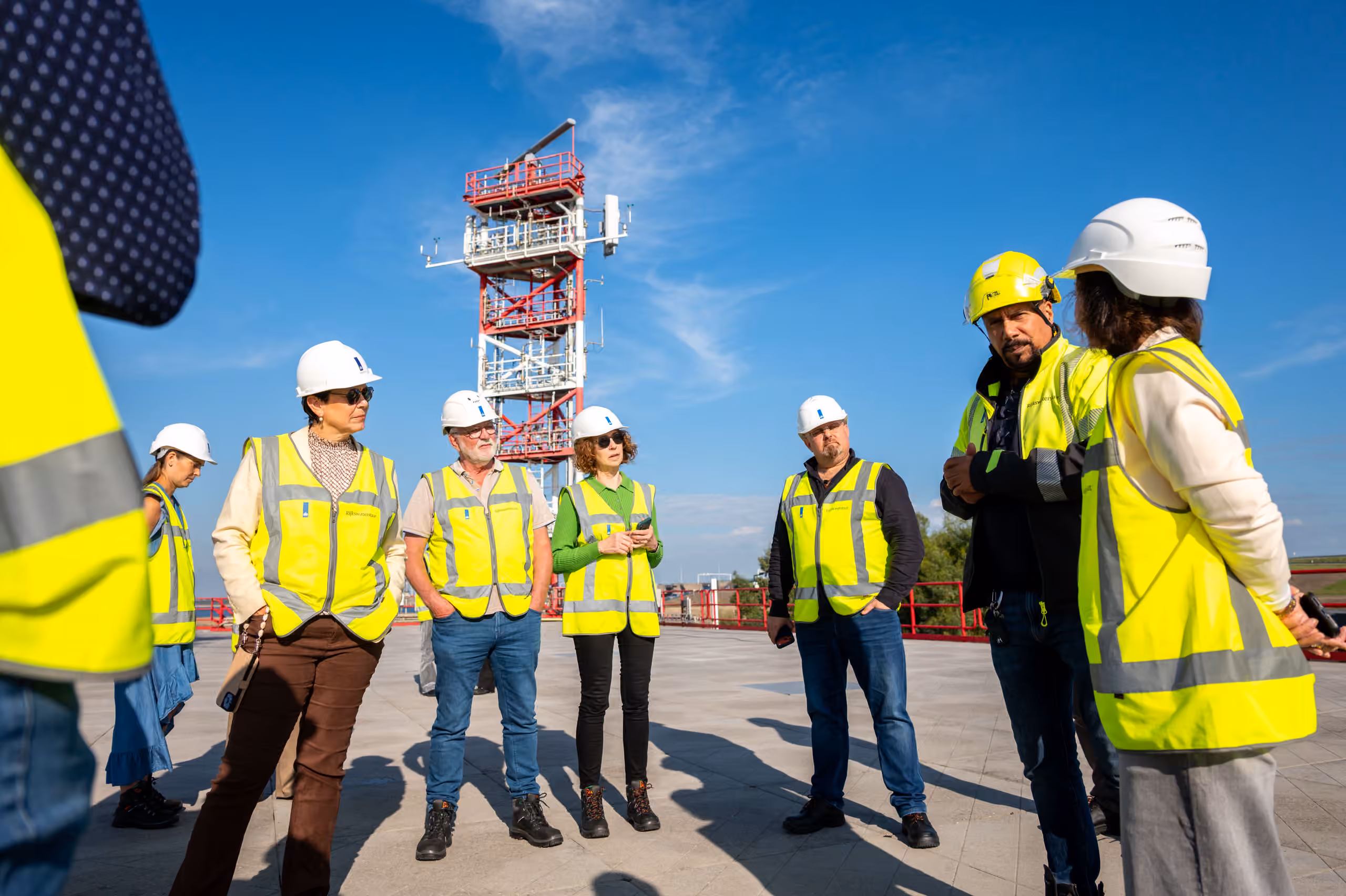 Een groep bezoekers met veiligheidsvesten en helmen krijgt buiten uitleg bij de testopstelling van het Offshore Expertise Centrum. Op de achtergrond staat een hoge rood-witte mast met meetapparatuur.