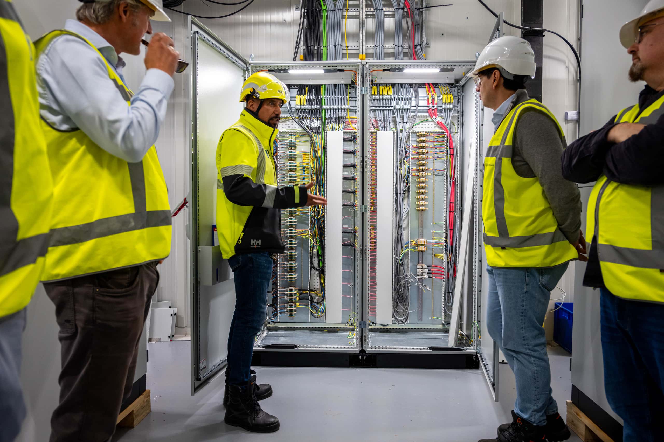 Bezoekers met veiligheidsvesten en helmen krijgen uitleg bij een geopende schakelkast met bekabeling en technische apparatuur in het Offshore Expertise Centrum.